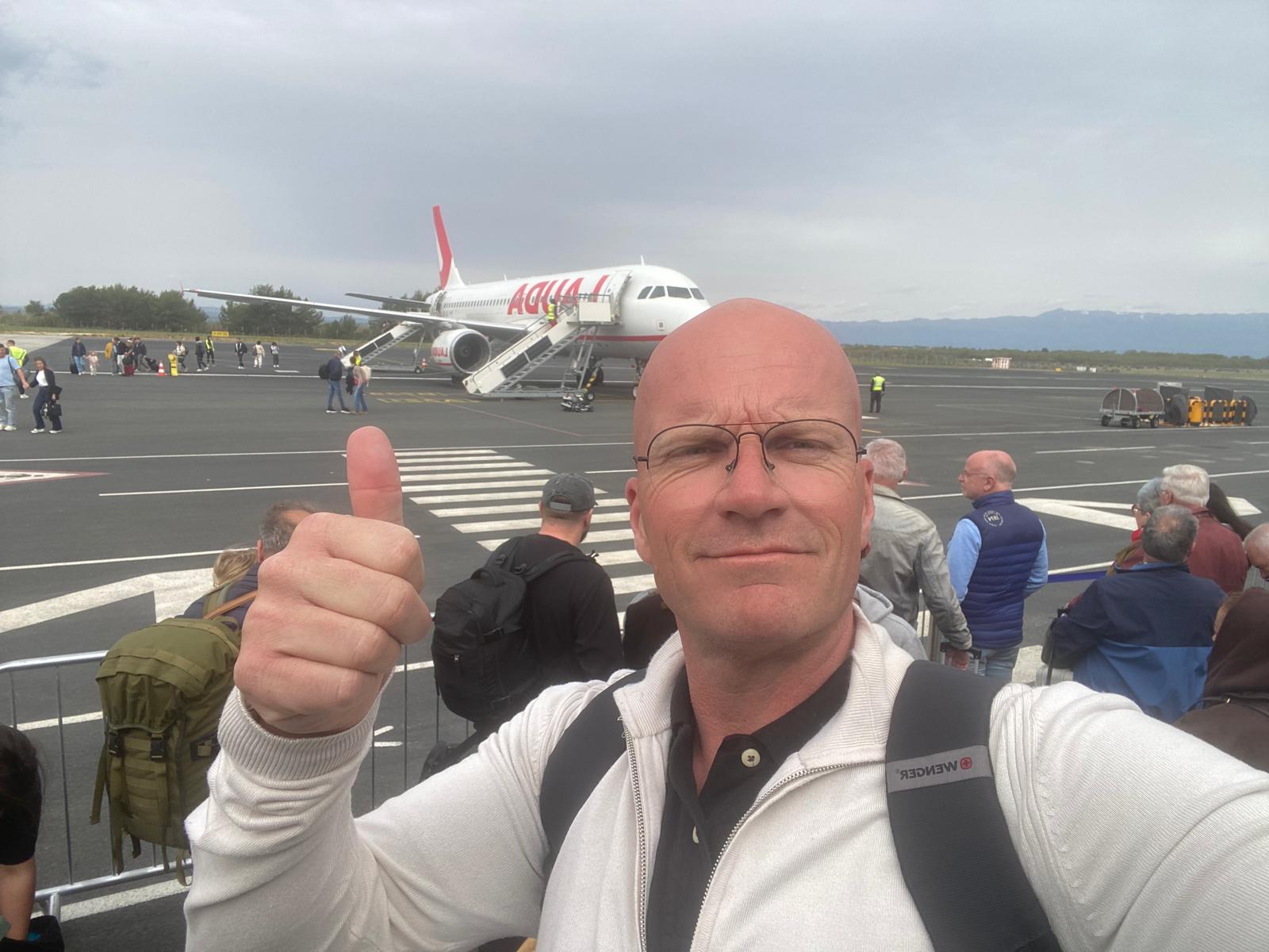 Joost van Beers geeft een duim omhoog op de luchthaven met een vliegtuig op de achtergrond